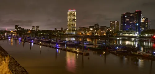 London view from across the thames from imperial wharf at night 2 The image shows an urban landscape view of London from across the River Thames near Imperial Wharf at night in early autumn. The riverbank is lined with boats, their lights reflecting off the water, creating a vibrant scene. Modern residential and commercial buildings dominate the skyline, with the distinctive cylindrical tower of Imperial Wharf standing out among the structures. The setting is in the United Kingdom, and the warm glow from the building lights and boat moorings enhances the atmosphere of this riverside area. The photograph captures the essence of London’s urban development along the River Thames and highlights the lively presence of boats and contemporary architecture, representative of this iconic riverside district.